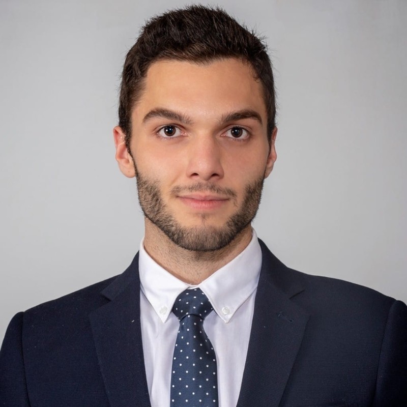 Portrait of Alhussein Jamil, a software engineer with short dark hair, wearing a navy suit, white shirt, and patterned blue tie, photographed against a neutral grey background.
